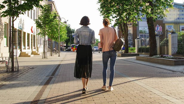 Two women enjoy a sunny walk down a lively urban street lined with buildings and trees.